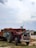 A red tractor with decorative patterns is attached to a loaded trailer stacked high with red bricks. The scene appears to be set outdoors on a cloudy day, with buildings and a grassy area in the background. The tractor has colorful artwork typical of local designs.