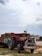 A red tractor with decorative patterns is attached to a loaded trailer stacked high with red bricks. The scene appears to be set outdoors on a cloudy day, with buildings and a grassy area in the background. The tractor has colorful artwork typical of local designs.