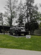 A military armored vehicle is displayed on a concrete platform surrounded by green grass. Tall trees with dense foliage form a backdrop, creating a natural setting. A small sign in front of the vehicle contains text indicating its type and purpose.