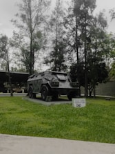 A military armored vehicle is displayed on a concrete platform surrounded by green grass. Tall trees with dense foliage form a backdrop, creating a natural setting. A small sign in front of the vehicle contains text indicating its type and purpose.