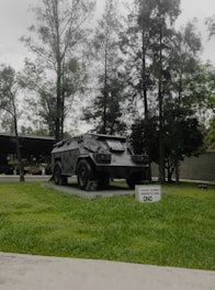 A military armored vehicle is displayed on a concrete platform surrounded by green grass. Tall trees with dense foliage form a backdrop, creating a natural setting. A small sign in front of the vehicle contains text indicating its type and purpose.