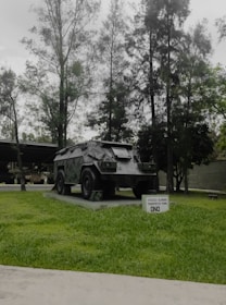 A military armored vehicle is displayed on a concrete platform surrounded by green grass. Tall trees with dense foliage form a backdrop, creating a natural setting. A small sign in front of the vehicle contains text indicating its type and purpose.