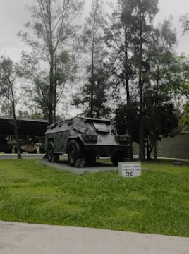 A military armored vehicle is displayed on a concrete platform surrounded by green grass. Tall trees with dense foliage form a backdrop, creating a natural setting. A small sign in front of the vehicle contains text indicating its type and purpose.