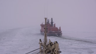 A large icebreaker ship is navigating through a frozen sea. The ship is painted in a vivid red color and appears to be breaking through the ice, leaving a path in its wake. The sea and sky blend together in shades of gray and white, with a foggy atmosphere. The foremast of another ship appears in the foreground.