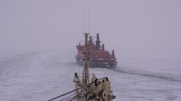 A large icebreaker ship is navigating through a frozen sea. The ship is painted in a vivid red color and appears to be breaking through the ice, leaving a path in its wake. The sea and sky blend together in shades of gray and white, with a foggy atmosphere. The foremast of another ship appears in the foreground.