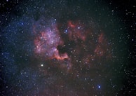 A vast expanse of the universe featuring countless stars scattered across the darkness of space. Intricate patterns of red and purple nebulous clouds form a visually striking region, illuminated by the surrounding starlight. Some areas exhibit denser star clusters, adding depth and complexity to the cosmic scene.