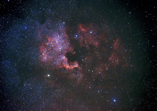 A vast expanse of the universe featuring countless stars scattered across the darkness of space. Intricate patterns of red and purple nebulous clouds form a visually striking region, illuminated by the surrounding starlight. Some areas exhibit denser star clusters, adding depth and complexity to the cosmic scene.