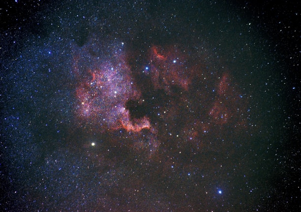 A vast expanse of the universe featuring countless stars scattered across the darkness of space. Intricate patterns of red and purple nebulous clouds form a visually striking region, illuminated by the surrounding starlight. Some areas exhibit denser star clusters, adding depth and complexity to the cosmic scene.