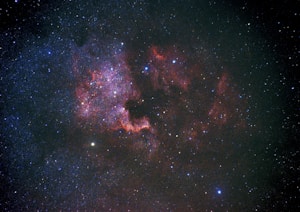 A vast expanse of the universe featuring countless stars scattered across the darkness of space. Intricate patterns of red and purple nebulous clouds form a visually striking region, illuminated by the surrounding starlight. Some areas exhibit denser star clusters, adding depth and complexity to the cosmic scene.