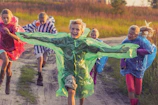 A group of children wearing clay heart outfits running barefoot on green grass under a sunny sky.
