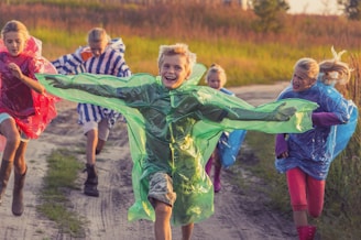 A group of children are running joyfully down a dirt path. They are wearing colorful rain ponchos - green, red, blue, and striped. The weather appears to be sunny, and there is lush grassy vegetation on either side of the path.