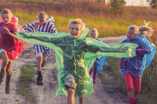 A group of children are running joyfully down a dirt path. They are wearing colorful rain ponchos - green, red, blue, and striped. The weather appears to be sunny, and there is lush grassy vegetation on either side of the path.