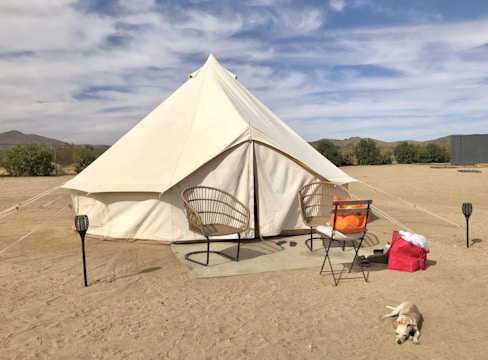 A white canvas tent is set up on a sandy surface in an arid landscape. Two wicker chairs and a folding chair with an orange cushion are placed in front of the tent. Nearby, a pink bag lies on the ground, and a small dog is resting peacefully in the sun. The background features sparse vegetation and distant hills under a partly cloudy sky.