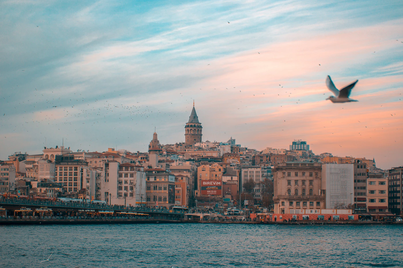 View of the Galata Tower and Istanbul skyline with mosques and minarets visible in the background and the Bosphorus waterway reflecting the soft sky in the foreground with boats on the water