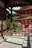 A traditional Japanese temple with wooden architecture is visible, featuring a large hanging bell and intricate red pagoda-style structures. Lush green vegetation surrounds the area, and sunlight casts shadows and contributes to the tranquil ambiance.