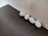 Four white eggs are arranged in a straight line against a light-colored wall. The eggs rest on a dark wooden surface, creating a contrast between the white of the eggs and the dark texture of the table. The lighting is soft, casting gentle shadows around the eggs.