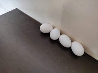 Four white eggs are arranged in a straight line against a light-colored wall. The eggs rest on a dark wooden surface, creating a contrast between the white of the eggs and the dark texture of the table. The lighting is soft, casting gentle shadows around the eggs.