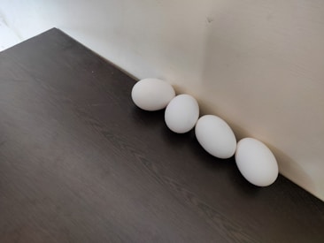 Four white eggs are arranged in a straight line against a light-colored wall. The eggs rest on a dark wooden surface, creating a contrast between the white of the eggs and the dark texture of the table. The lighting is soft, casting gentle shadows around the eggs.