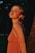 A young woman with light hair is captured in a warm, golden light. She gazes to the side, wearing a sleeveless, peach-toned dress. The background is a dark, blurred forest setting.