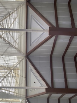 A geometric architectural design featuring triangular and rectangular patterns formed by metal beams and panels. The structures create a symmetrical and modern aesthetic with a combination of horizontal slats and angled beams.