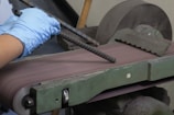A pair of hands wearing blue gloves is holding a piece of rebar against a belt sander. The belt sander is on and smoothing the surface of the rebar. The machine is green with visible wear and is situated in a workshop setting.