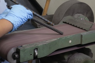 A pair of hands wearing blue gloves is holding a piece of rebar against a belt sander. The belt sander is on and smoothing the surface of the rebar. The machine is green with visible wear and is situated in a workshop setting.