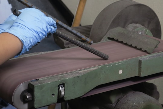 A pair of hands wearing blue gloves is holding a piece of rebar against a belt sander. The belt sander is on and smoothing the surface of the rebar. The machine is green with visible wear and is situated in a workshop setting.