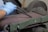 A pair of hands wearing blue gloves is holding a piece of rebar against a belt sander. The belt sander is on and smoothing the surface of the rebar. The machine is green with visible wear and is situated in a workshop setting.