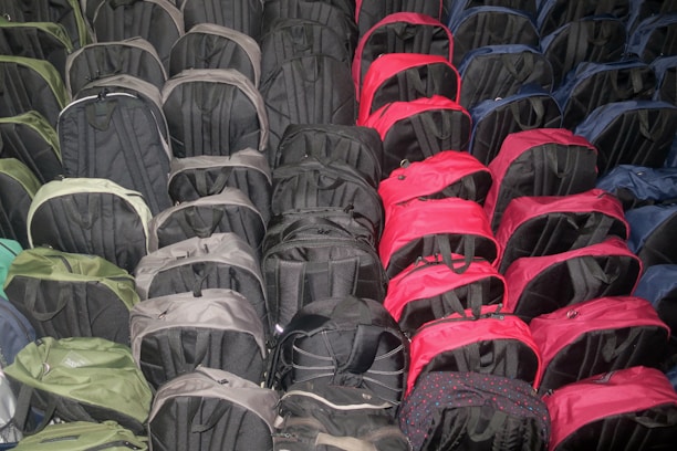 A large collection of backpacks is arranged in neat rows. The backpacks are mainly black, with some appearing in red, green, and blue. They are closely packed together, occupying the entire visible area.