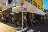 A street corner featuring a small local shop with awnings, displaying umbrellas and assorted goods on the sidewalk. The building has a yellow and gray facade. Pedestrians are browsing the items, and a red bicycle is parked nearby. There are road signs and a crosswalk marking in front.