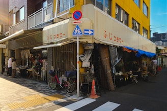 A street corner featuring a small local shop with awnings, displaying umbrellas and assorted goods on the sidewalk. The building has a yellow and gray facade. Pedestrians are browsing the items, and a red bicycle is parked nearby. There are road signs and a crosswalk marking in front.