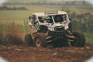 A rugged off-road vehicle is speeding through a dirt track, kicking up a spray of mud and debris. The background features a blurred landscape of fields or countryside. The vehicle has large, robust tires and a reinforced frame, indicating it's built for tough terrains.