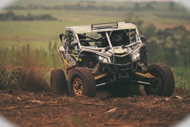 A rugged off-road vehicle is speeding through a dirt track, kicking up a spray of mud and debris. The background features a blurred landscape of fields or countryside. The vehicle has large, robust tires and a reinforced frame, indicating it's built for tough terrains.