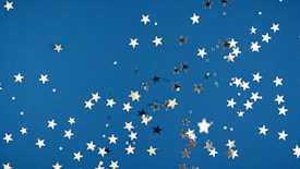 A collection of small, shiny star-shaped confetti scattered across a vibrant blue background. The stars vary in size and appear in gold, silver, and black, adding a sense of sparkle and celebration.