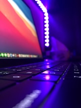 Cinematic close-up of a glowing neon purple and pink light reflecting on a sleek modern desk with a laptop and camera gear.