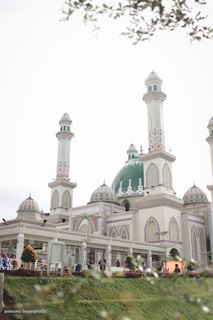 A beautiful mosque with multiple minarets and a large green dome. The building features intricate patterns and decorations, with well-maintained gardens surrounding it. Several people are seen walking around the premises, and the sky is overcast, contributing to a serene ambiance.