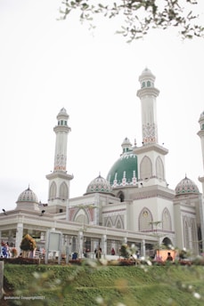 A beautiful mosque with multiple minarets and a large green dome. The building features intricate patterns and decorations, with well-maintained gardens surrounding it. Several people are seen walking around the premises, and the sky is overcast, contributing to a serene ambiance.