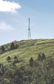 A tall communication tower stands prominently on a green hillside under a clear blue sky. The hill is covered with scattered trees and shrubs, and a small white vehicle is visible on a winding road. A large red and white sign is partially visible on the left side of the hill.