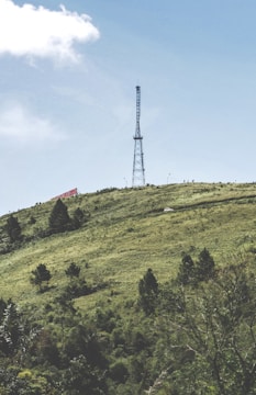 A tall communication tower stands prominently on a green hillside under a clear blue sky. The hill is covered with scattered trees and shrubs, and a small white vehicle is visible on a winding road. A large red and white sign is partially visible on the left side of the hill.