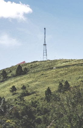 A tall communication tower stands prominently on a green hillside under a clear blue sky. The hill is covered with scattered trees and shrubs, and a small white vehicle is visible on a winding road. A large red and white sign is partially visible on the left side of the hill.