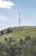 A tall communication tower stands prominently on a green hillside under a clear blue sky. The hill is covered with scattered trees and shrubs, and a small white vehicle is visible on a winding road. A large red and white sign is partially visible on the left side of the hill.