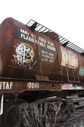 A rusted railway tanker, likely used for transporting flammable liquids, is situated on train tracks. The tanker is covered with graffiti and displays a sign indicating its maximum load capacity of 44,500 liters or 44.5 tonnes. There are steel ladders and walkways attached to the tanker, and dry vegetation can be seen in the foreground.