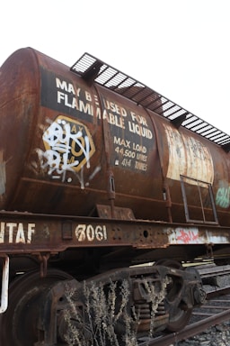 A rusted railway tanker, likely used for transporting flammable liquids, is situated on train tracks. The tanker is covered with graffiti and displays a sign indicating its maximum load capacity of 44,500 liters or 44.5 tonnes. There are steel ladders and walkways attached to the tanker, and dry vegetation can be seen in the foreground.
