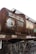 A rusted railway tanker, likely used for transporting flammable liquids, is situated on train tracks. The tanker is covered with graffiti and displays a sign indicating its maximum load capacity of 44,500 liters or 44.5 tonnes. There are steel ladders and walkways attached to the tanker, and dry vegetation can be seen in the foreground.