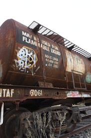 A rusted railway tanker, likely used for transporting flammable liquids, is situated on train tracks. The tanker is covered with graffiti and displays a sign indicating its maximum load capacity of 44,500 liters or 44.5 tonnes. There are steel ladders and walkways attached to the tanker, and dry vegetation can be seen in the foreground.
