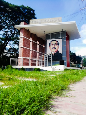 A modern architectural structure features a large portrait on the facade. The building incorporates brick and metal elements and is surrounded by lush greenery. The sky above is bright and clear, with light utility wiring visible in the distance.