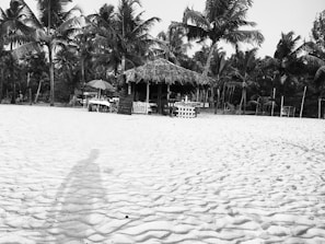 A sandy beach with visible footprints leads to a thatched hut surrounded by palm trees. In front of the hut, there are tables and chairs with umbrellas providing shade. The setting appears relaxed and tropical, offering a sense of retreat and leisure.