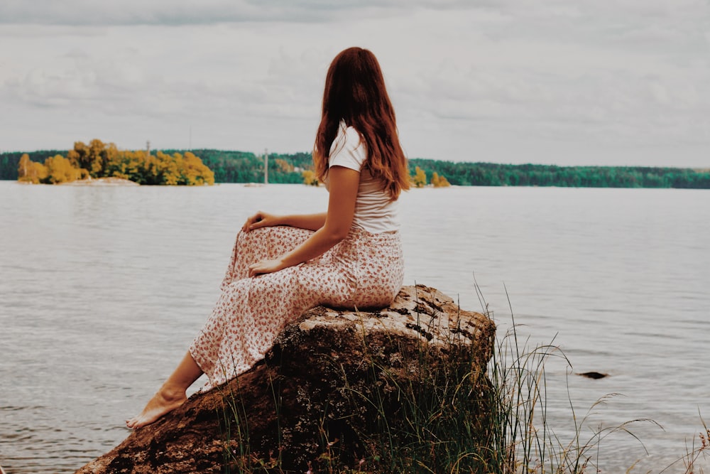 woman in white dress sitting on brown rock near body of water during daytime