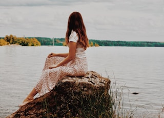 woman in white dress sitting on brown rock near body of water during daytime