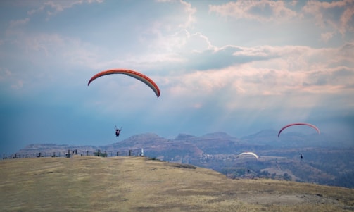 Paragliders soar over a vast, open landscape with rolling hills and a dramatic sky filled with clouds and sunlight rays.